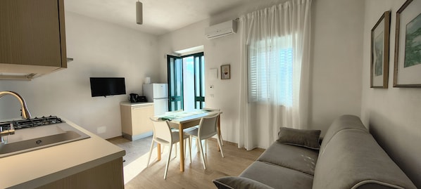 Une cuisine moderne équipée d’une cuisinière intégrée, un coin repas avec une table et des chaises, un canapé gris et un téléviseur fixé au mur.