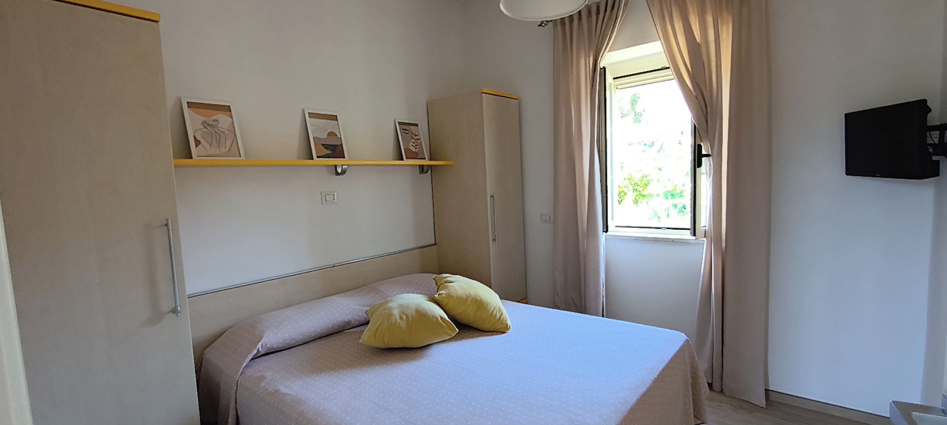 Ein Schlafzimmer mit Bett, zwei Kissen, einem Regal mit Kunstwerken, einem Kleiderschrank, einem Fenster mit Vorhängen und einem Wand-Fernseher.