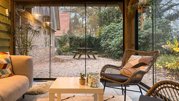 A cozy room with a sofa, a wicker chair, and a small table. There is a large window with a view of a garden and a brick building.
