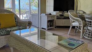 Una sala de estar con una mesa de centro de vidrio, muebles de mimbre, un televisor y una estantería.