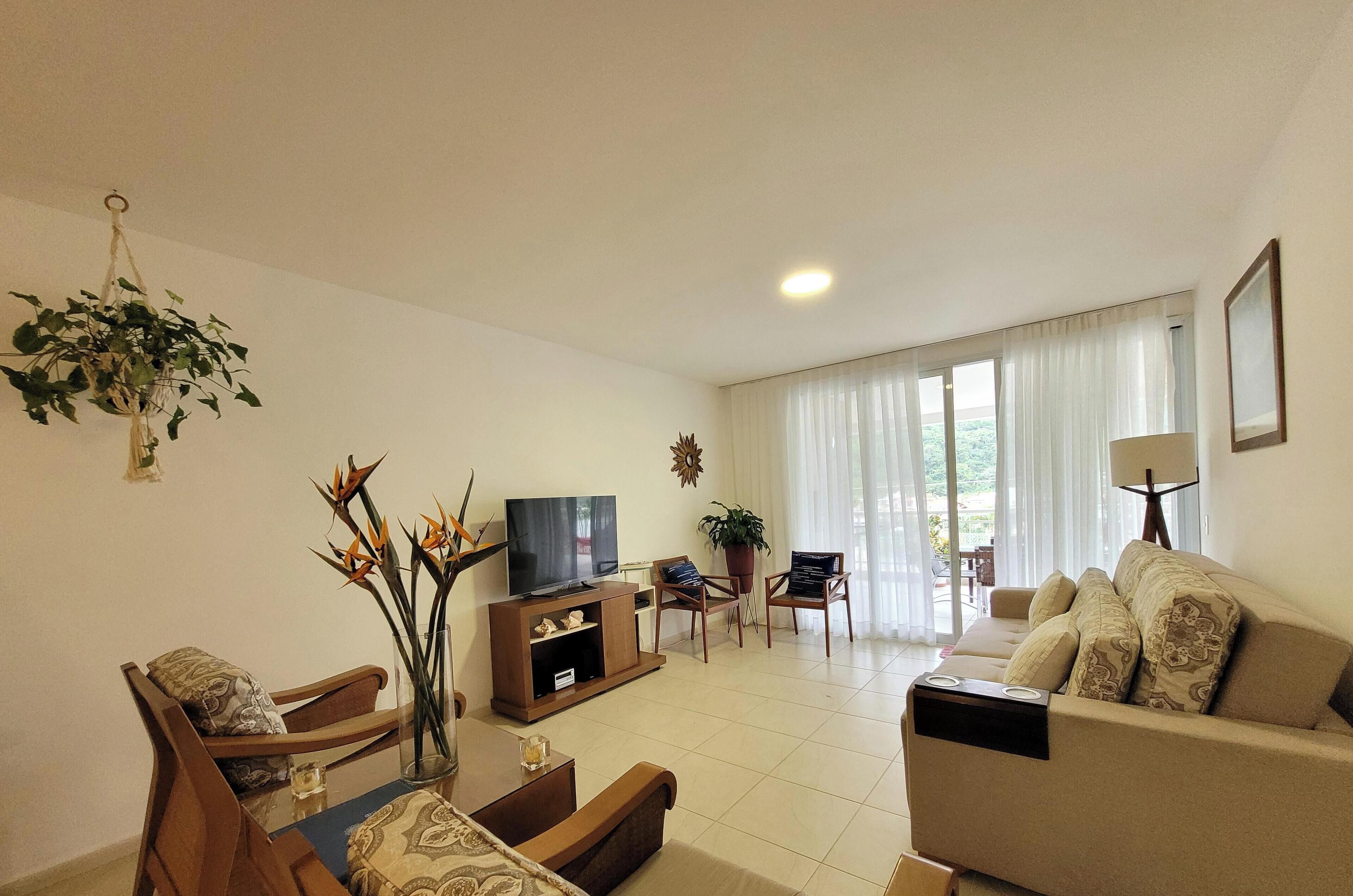 Una sala de estar con un sofá, televisor, sillas de madera, una mesa de comedor y una planta colgante.