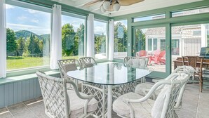 A sunlit room with a glass table surrounded by wicker chairs, large windows, and a view of greenery.