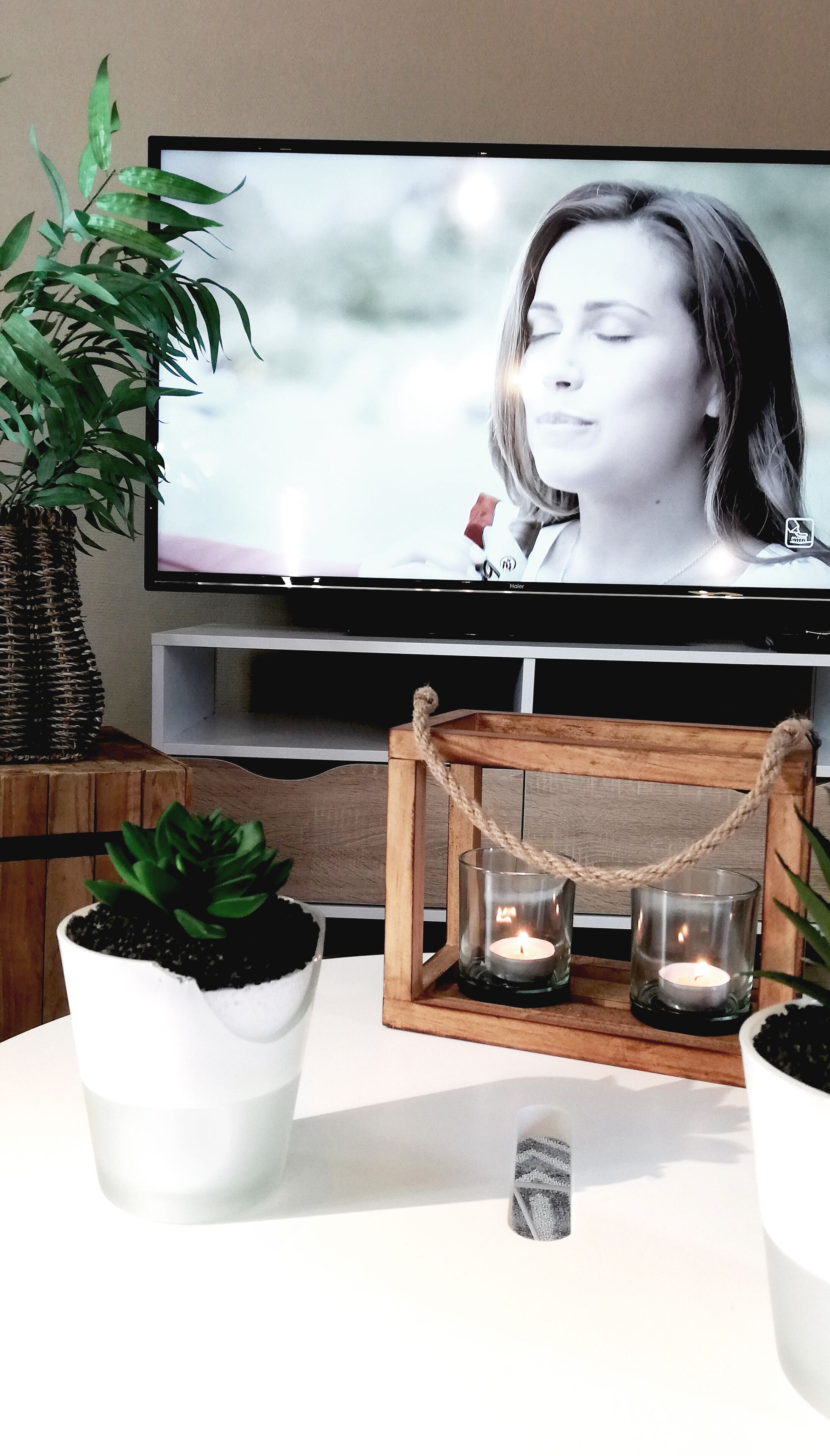 Een televisie toont een persoon, een potplant en een kandelaar met twee kaarsen op een tafel.