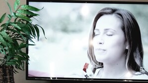 A television displaying a person, a potted plant, and a candle holder with two candles on a table.