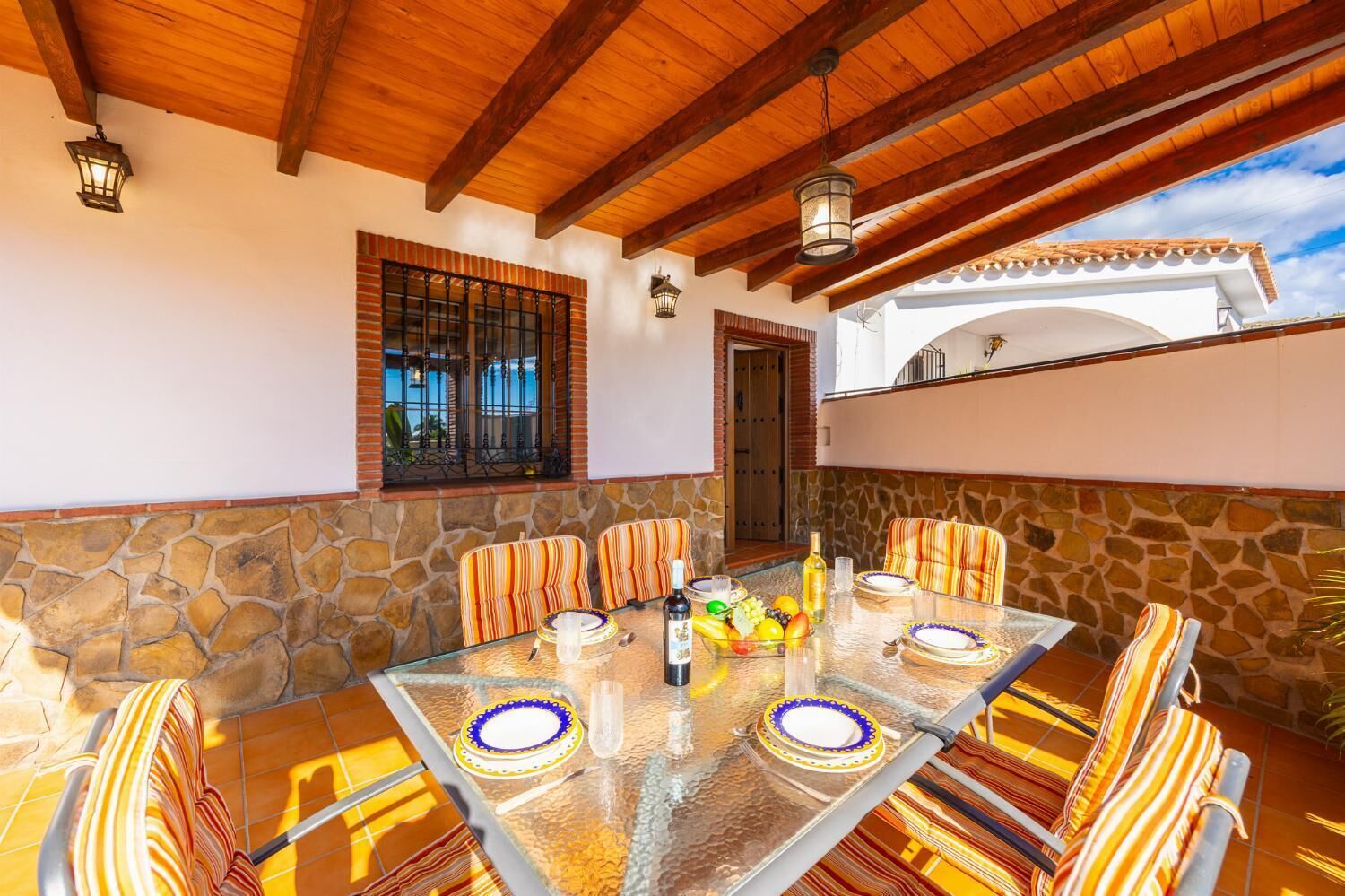 Un patio al aire libre con una mesa de vidrio, sillas a rayas, un muro de piedra y un techo de madera.
