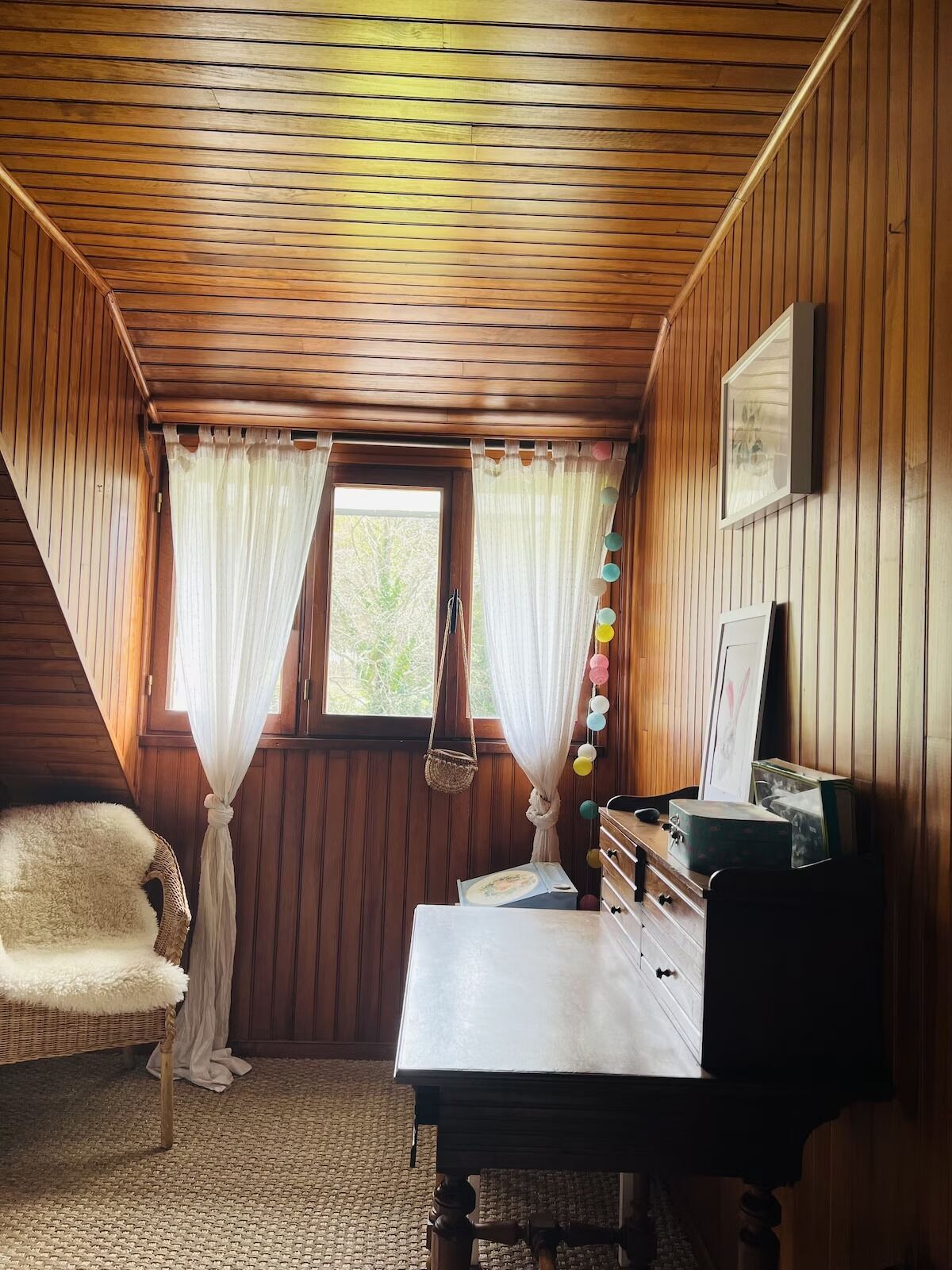 Une pièce chaleureuse avec des boiseries, un bureau doté de tiroirs, un fauteuil en osier et une fenêtre avec des rideaux.