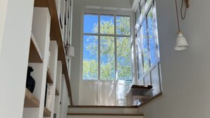 Eine Treppe mit Holzboden und weißen Wänden führt zu einem hellen Raum mit einem großen Fenster und einer Pendelleuchte.
