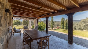 Un patio techado al aire libre con una mesa y sillas de madera, paredes de piedra y vistas al paisaje.