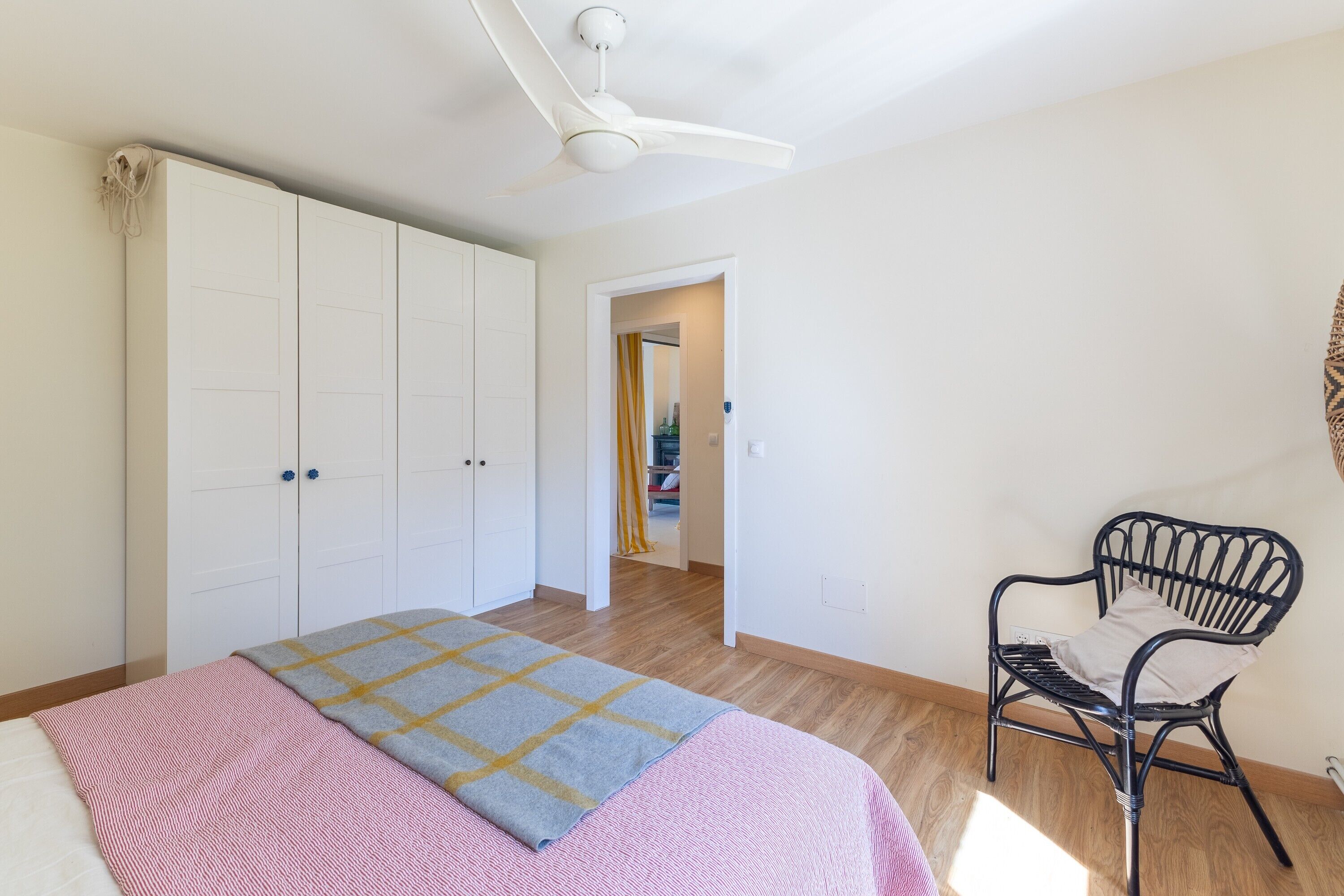 Un dormitorio con cama, ventilador de techo, armario, una silla y un acceso a otra habitación.