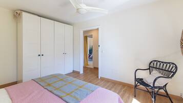 Un dormitorio con cama, ventilador de techo, armario, una silla y un acceso a otra habitación.