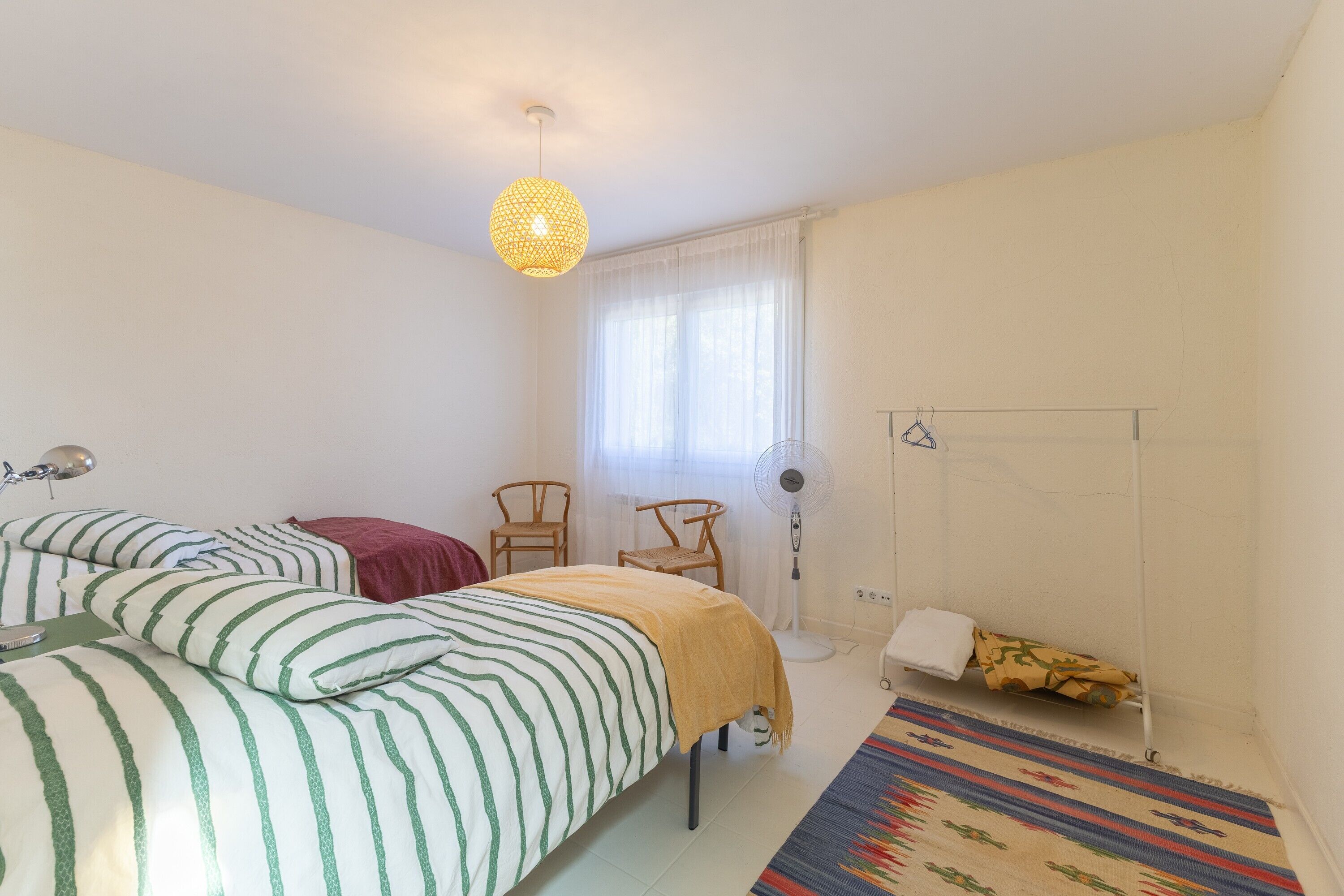 Un dormitorio con dos camas, un ventilador, una ventana con cortinas y una alfombra estampada.