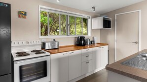 A modern kitchen with white cabinets, a stove, a window with a view of trees, and a countertop with a coffee maker. - Treetop Break - Waipu Cove Holiday Home (Waipu Cove)