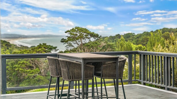 A balcony with a table and chairs, overlooking a scenic view of a beach and forest. - Treetop Break - Waipu Cove Holiday Home (Waipu Cove)