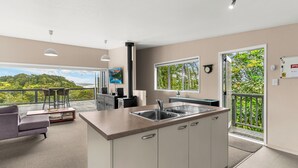 A modern kitchen with a large island, a dining area with a sofa and chairs, and a balcony with a view of greenery. - Treetop Break - Waipu Cove Holiday Home (Waipu Cove)