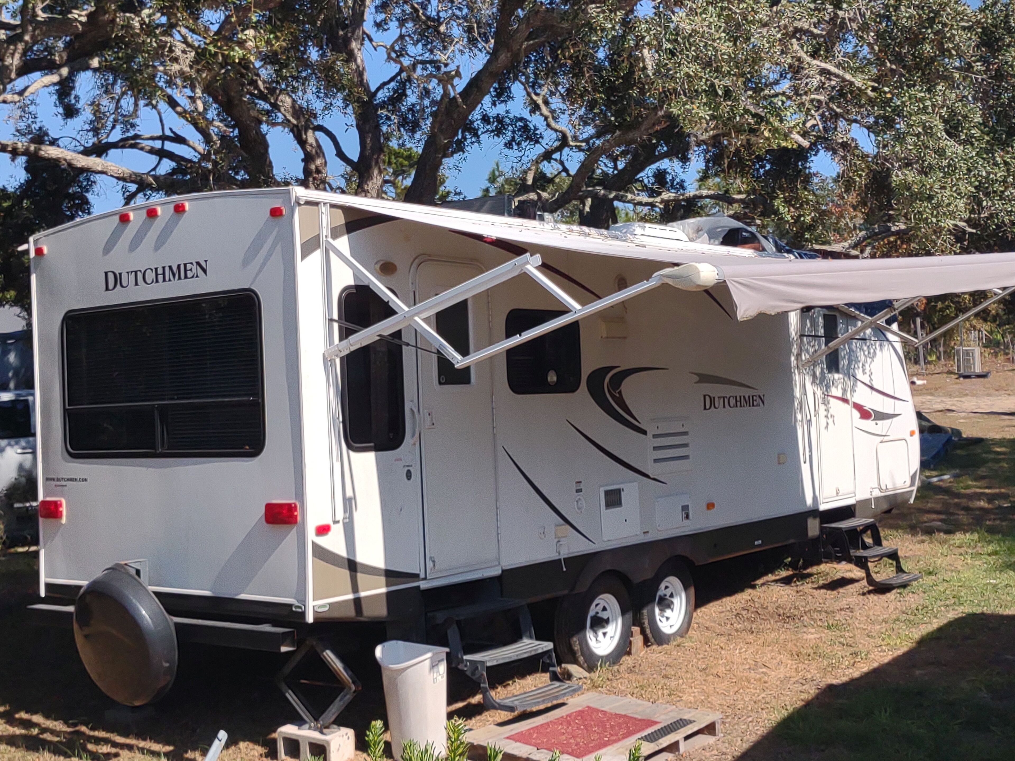 A white RV with the brand name 'DUTCHMEN' on the side, featuring a covered awning and a small step-up platform.