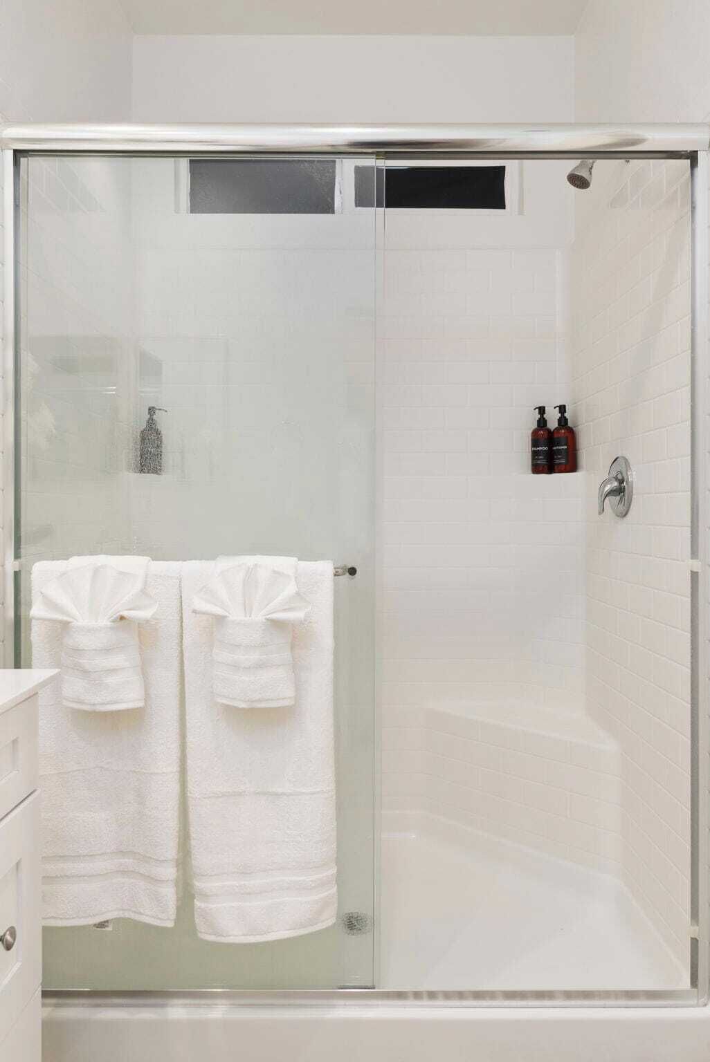 A glass-enclosed shower stall with a white tiled wall, a glass door, and two white towels hanging on a towel rack.