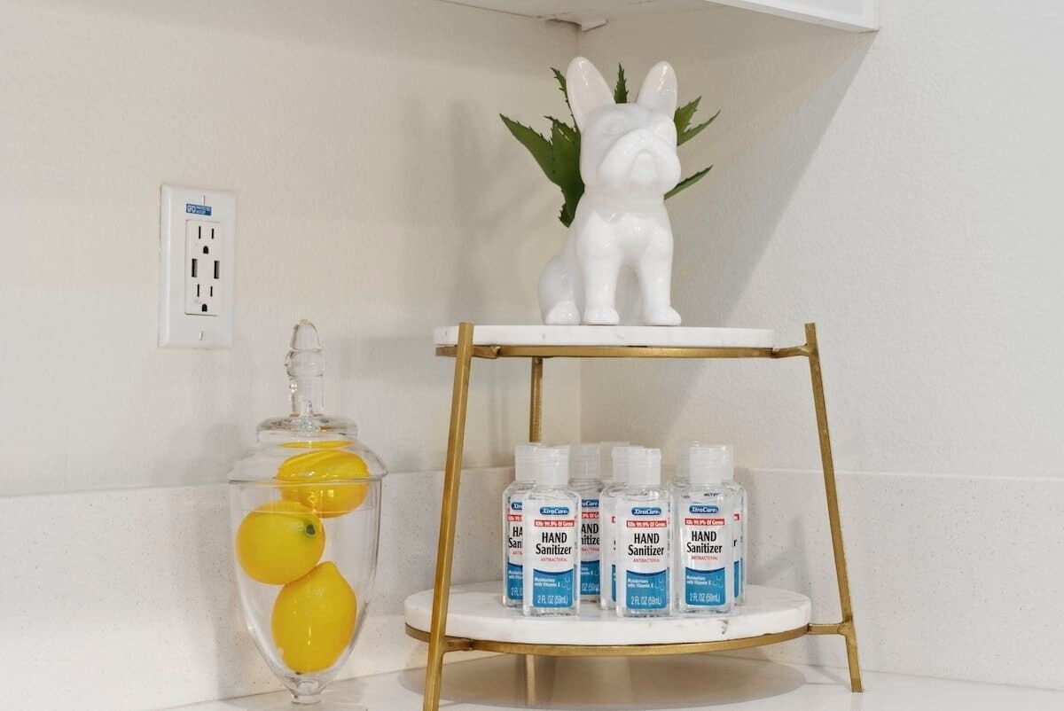 Een badkamer met een witte muur, een witte keramieken konijnenbeeldje, een glazen vaas met citroenen en drie flesjes handdesinfectiemiddel op een gouden plank.