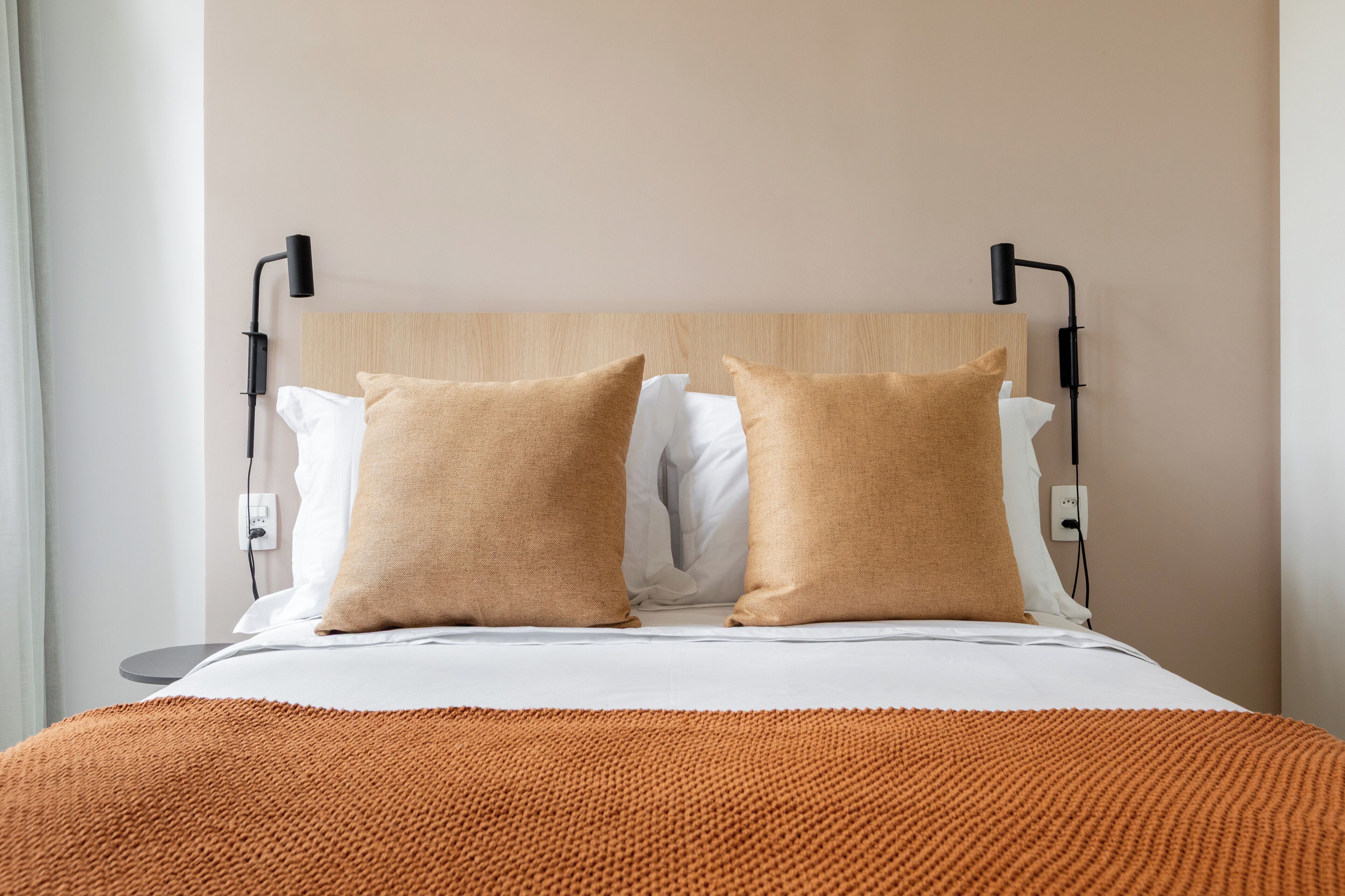 En snyggt bäddad säng med en sänggavel i trä, två beige kuddar och en orange stickad pläd.