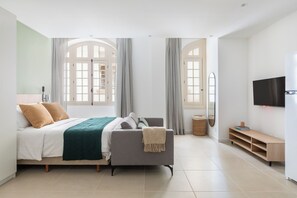 Un dormitorio moderno con una cama, un sofá gris, un televisor y un ventanal con cortinas.