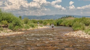 A river with two people fishing, surrounded by greenery and mountains in the background. - Charming Riverside Ranch Retreat with Horse Pasture Views (Bondurant)