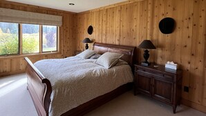 A bedroom with a wooden paneled wall, a large bed with a headboard, two bedside tables with lamps, and a window with a view of greenery. - Private Pond-Side Manor on a Working Wyoming Ranch (Bondurant)