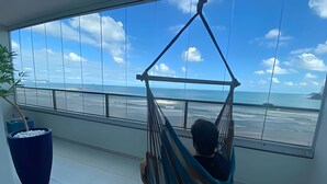 Una habitación con un ventanal grande con vistas al océano, una silla hamaca, una planta en maceta y un cubo de basura azul.