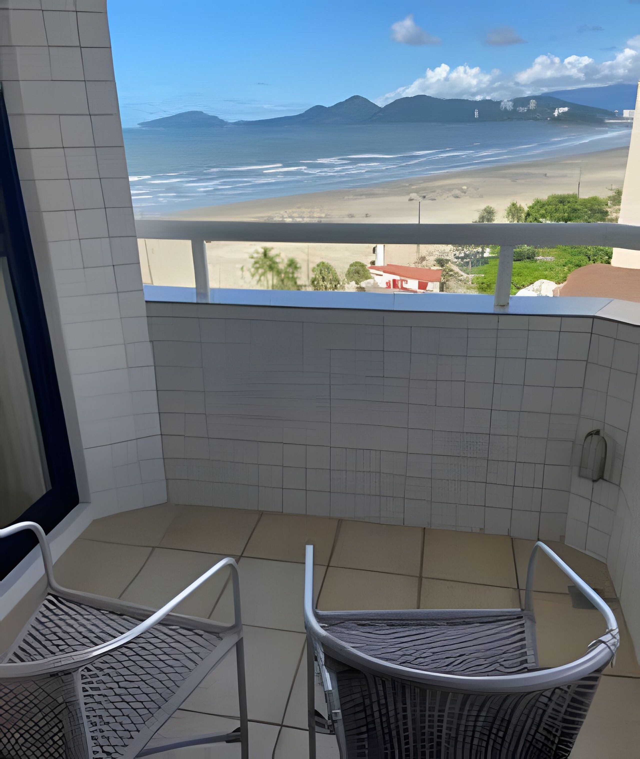 A balcony with two wicker chairs, a tiled floor, and a view of the beach and mountains.