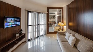 A modern living room with a flat-screen TV mounted on a wooden wall, a sofa with cushions, a lamp on a side table, and a sliding glass door leading to a kitchen area. - SHB - Elegant apartment with sea view in Gonzaga (Santos)