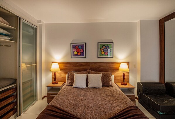 Une chambre à coucher avec un grand lit, deux tables de chevet avec des lampes, une armoire et deux œuvres d’art encadrées accrochées au mur.