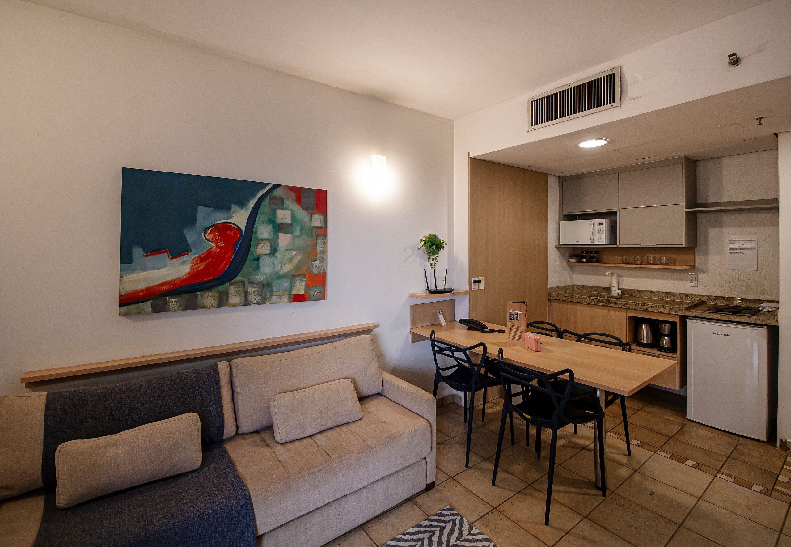 A modern living room with a sofa, a dining table with chairs, a kitchen area with cabinets and appliances, and a large abstract painting on the wall.