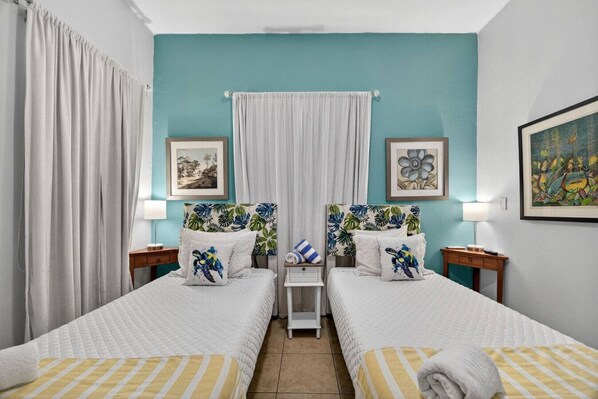 Une chambre double avec deux lits, chacun recouvert d’une courtepointe blanche matelassée et agrémenté d’oreillers à rayures. On y trouve deux tables de chevet avec des lampes, une petite table avec une serviette et une fenêtre avec des rideaux blancs.