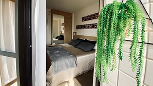 A bedroom with a bed, a bedside table, a hanging plant, and a wall with framed pictures. - SHB - Sea Glow: Balcony with Ocean View (Santos)