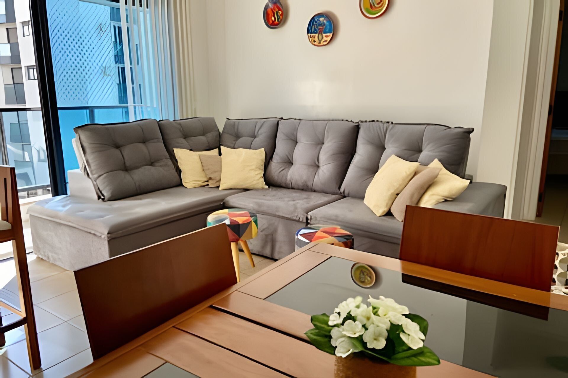 Een moderne woonkamer met een grijze hoekbank, een houten salontafel en een bijzettafeltje met een glazen blad en een bloemstuk.