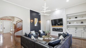 A modern living room with a dark leather sofa, a coffee table, and a flat-screen TV mounted on the wall. - 6BD Luxury Retreat w Backyard & Access Golf Course (Frisco)