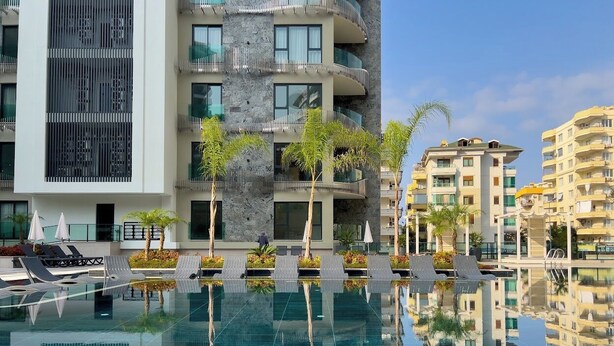 A modern hotel with a swimming pool, sun loungers, and a view of surrounding buildings. - Apartment 1+1 Qoople Excellence С49 With a terrace on the 1st line of the sea (Alanya)
