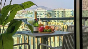 A balcony with a table set with a bottle of champagne, a fruit bowl, and a view of buildings and a mountain in the background. - Apartment 1+1 Qoople Excellence С49 With a terrace on the 1st line of the sea (Alanya)