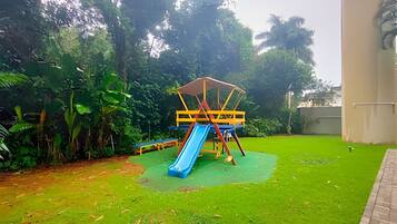 A playground with a slide and climbing structure, surrounded by greenery and a building.