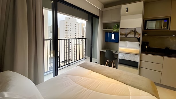 A hotel room with a bed, a desk, a kitchenette, and a view of buildings. - Studio near Sirio Libanes and Frei Caneca (São Paulo)