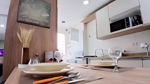 A modern kitchen with white cabinets, a microwave, and a dining table set with plates, cutlery, and a vase of dried flowers. - SHB - Wake Up with the Breeze: Charming Apartment in Embaré (Santos)