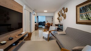 A modern living room with a sofa, TV, and wooden side table. There is a dining area with chairs and a table in the background. - SHB - Style and Comfort with Gourmet Area (Guarujá)