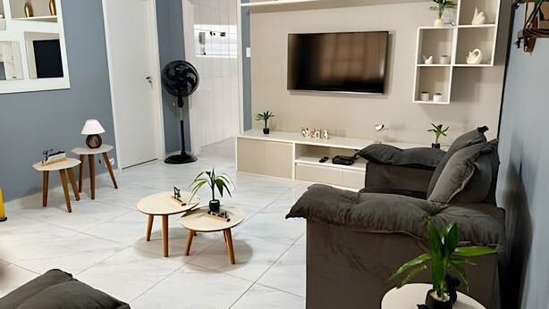 Un salon moderne avec un canapé gris, un téléviseur fixé au mur et un meuble de rangement blanc doté d’étagères. On y trouve deux petites tables rondes avec des lampes et un ventilateur sur pied.
