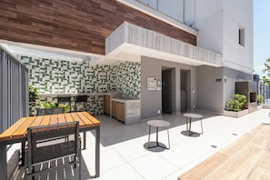 A modern outdoor dining area with a wooden table and chairs, a small kitchen area, and a seating space with two round tables. - Studio pet Friendly a 350m do Nubank Parque (São Paulo)