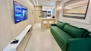 Un salon moderne avec un téléviseur à écran plat fixé au mur, un canapé vert, une console blanche et un coin cuisine à l’arrière-plan.