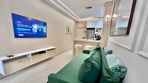 Un salon moderne avec un téléviseur à écran plat fixé au mur, un meuble de rangement blanc, un canapé vert avec des coussins, et un coin cuisine à l’arrière-plan.