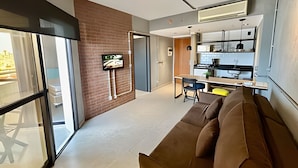 En moderne stue med en brun sofa, et tv monteret på en murstensvæg, og et køkkenområde med skabe og en bordplade.