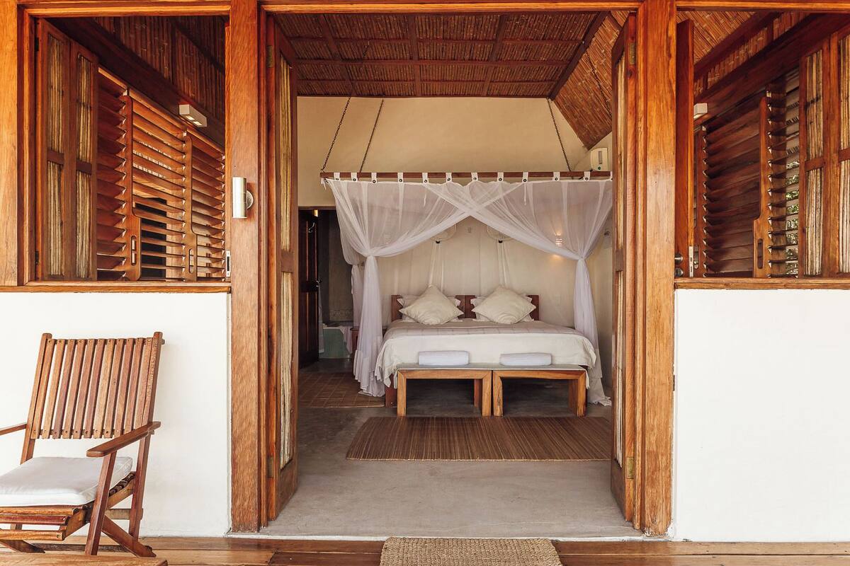 A wooden-framed room with a bed under a white canopy, a wooden chair, and a concrete floor.