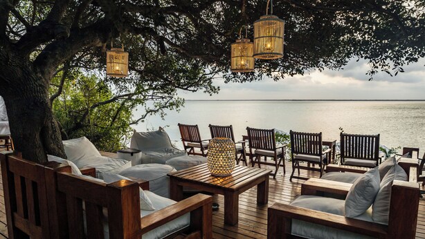 A wooden deck with outdoor furniture, including sofas and chairs, under a large tree with hanging lanterns. - Rio Azul Mozambique  (Bartolomeu Dias Point)