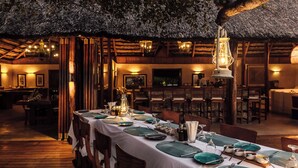 A restaurant with a thatched roof, wooden beams, and a long dining table set with green placemats, plates, and candles. - Rio Azul Mozambique  (Bartolomeu Dias Point)