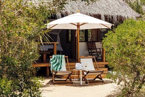 A wooden deck with two lounge chairs, a table with a glass of wine, and a thatched umbrella. - Rio Azul Mozambique  (Bartolomeu Dias Point)