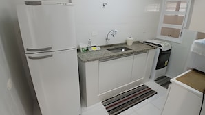 A kitchen with white cabinets, a granite countertop, a sink, a refrigerator, and a window with blinds. - SHB - Facing the Sea at Pitangueiras Beach (Guarujá)